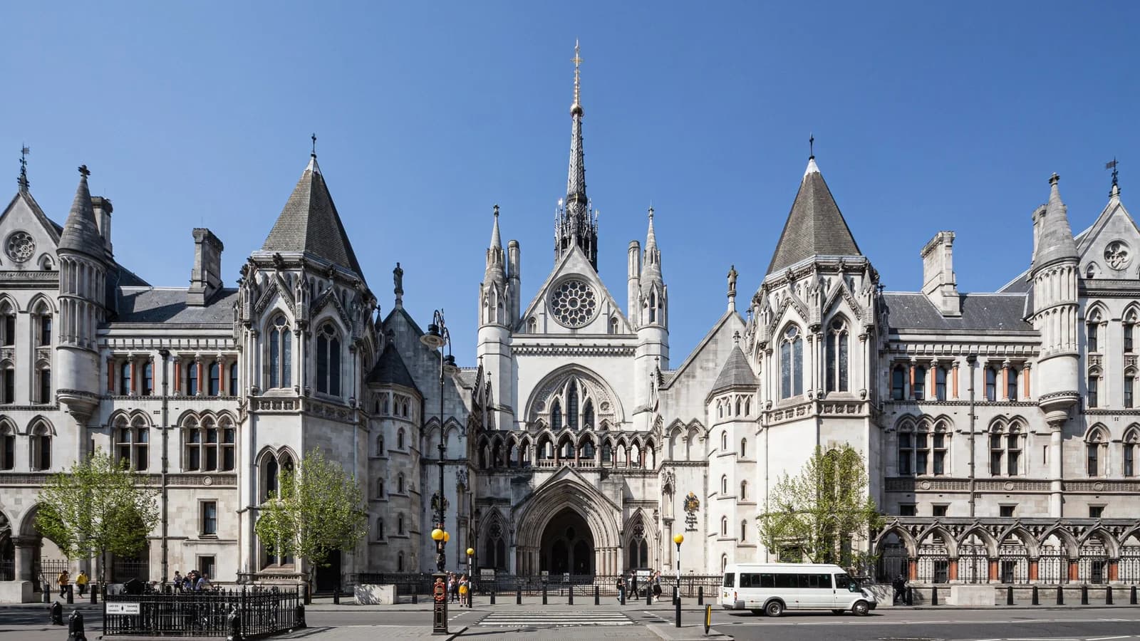 Hauptfassade der Royal Courts of Justice in London mit neugotischem Mittelportal, Rosettenfenster und Spitzturm unter blauem Himmel, davor die Straße