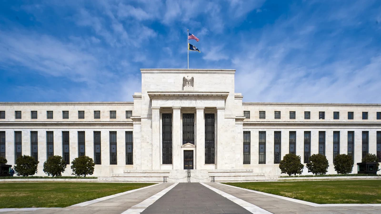 Frontansicht des Eccles Building der US-Notenbank Federal Reserve in Washington: Marmorfassade mit Säulenportal, Adler und wehender US-Flagge unter blauem Himmel
