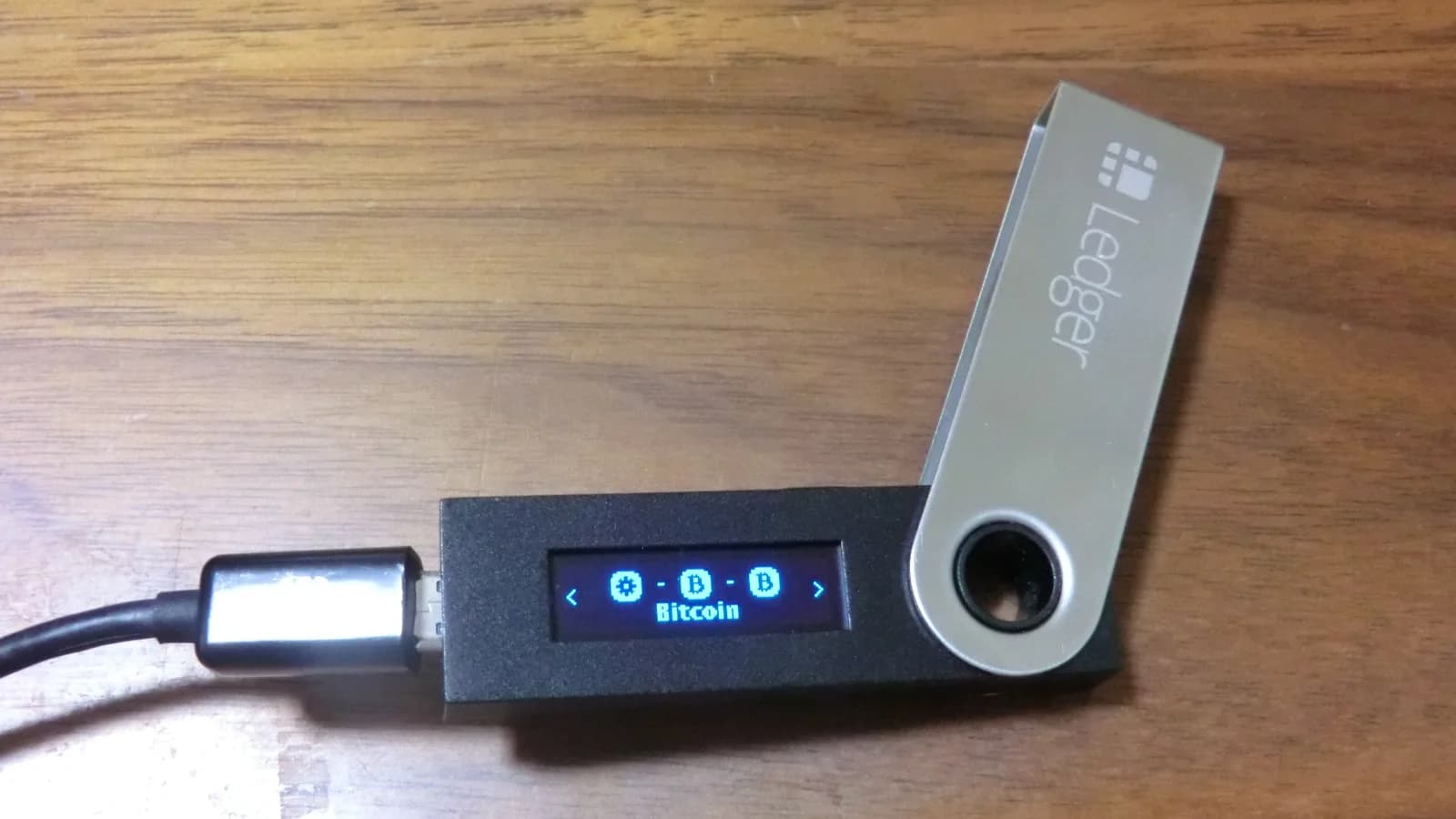 Ledger Nano S, per USB-Kabel angeschlossen, auf einem Holztisch. Auf dem OLED-Display steht „Bitcoin“, die Metallkappe ist aufgeklappt.