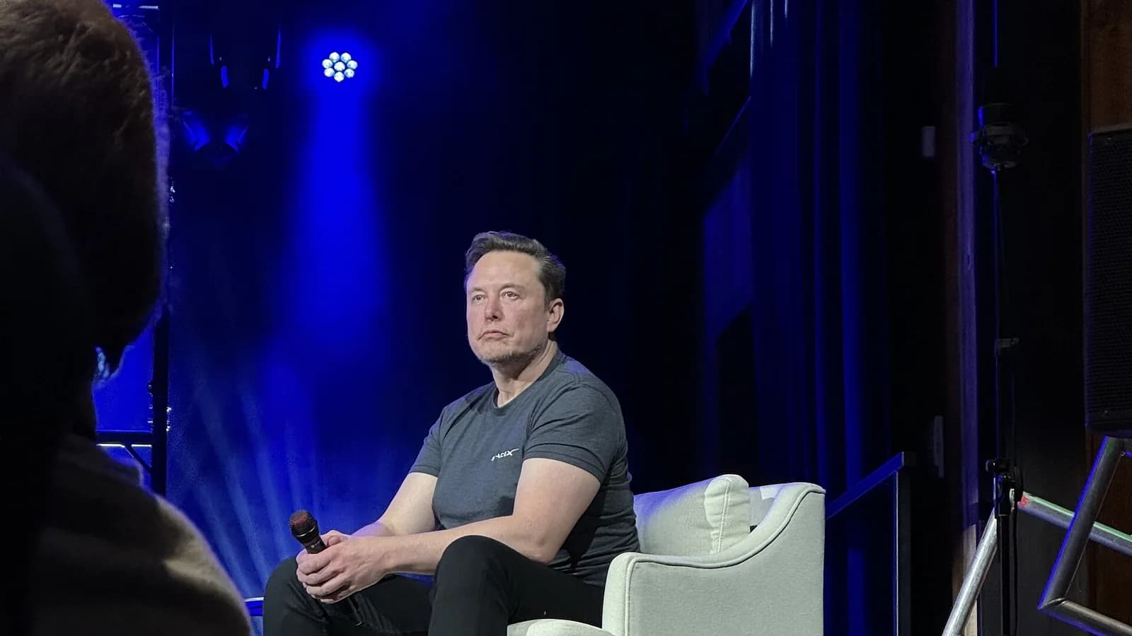 Elon Musk sitzt in dunklem T-Shirt und schwarzer Hose mit Mikrofon in der Hand auf einem hellen Sessel auf einer blau beleuchteten Bühne und blickt ins Publikum