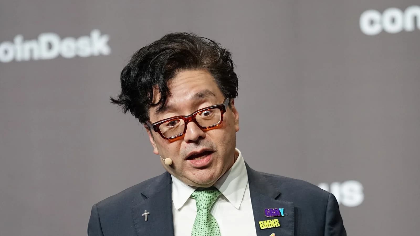 Tom Lee, Chairman von Bitmine, spricht auf der Bühne der Konferenz Consensus Hong Kong 2026, im dunklen Anzug mit grüner Krawatte, Headset und Presenter in der Hand